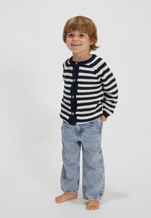 Marineblau und weiße gestreifte Strickjacke mit Holzknöpfen, kombiniert mit hellblauen, lässigen Jeans. Barfuß stehendes Kind lächelt.