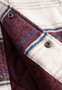 Țesătură de lână în carouri în nuanțe de alb, burdund și albastru, prezentând o căptușeală matlasată de culoare vin și detalii de închidere cu capse metalice.