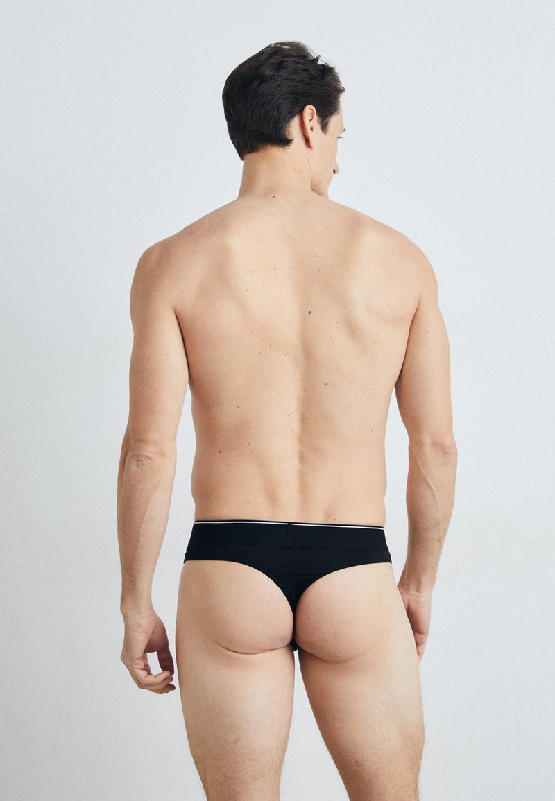 Schwarze Baumwoll-Tanga-Unterwäsche mit einem kontrastierenden Bund, minimaler Bedeckung und einer engen Passform, von hinten an einem Modell gezeigt.