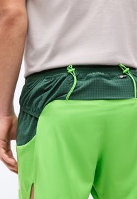 Άνδρας που φοράει στρωματωμένα πράσινα αθλητικά σορτς με ελαστική μέση και κορδόνια, συνδυασμένα με ανοιχτό γκρι πουκάμισο.