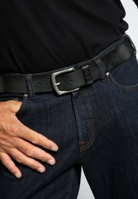 Sort læderbælte med en sølvmetal spænde, båret over mørke denim bukser. Bæltet har en struktureret overflade og flere justeringshuller.