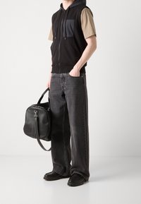 Muž v černé bezrukávové mikině přes béžové tričko, šedých širokých džínách, černých botách, držící černou koženou cestovní tašku.