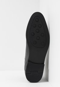 Svart lädersula med nio runda gummistift på framfoten, en texturerad häl, placerad vertikalt mot ett vitt hörn.