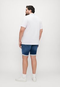 Camisa polo blanca, pantalones cortos de mezclilla azules con los puños enrollados, zapatillas blancas y calcetines blancos de canalé. Fondo liso.