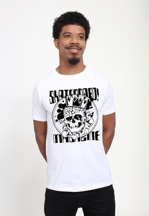 Mann mit kurzen lockigen Haaren, der ein weißes T-Shirt mit einem schwarzen Totenkopf und fettem Text trägt, steht vor einem einfarbigen Hintergrund.