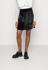 Gonna plissettata in velluto nero con una texture lucida e accenti scintillanti, indossata con scarpe nere lucide con suole spesse.
