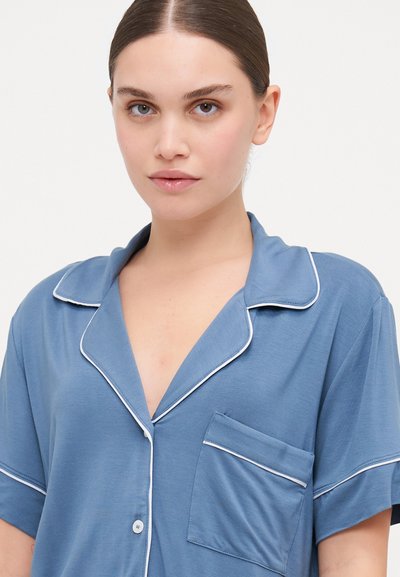 Camisa azul de manga curta com colarinho em notch, detalhes brancos e um bolso no peito. Feita de um tecido suave, apresenta um corte descontraído e textura lisa.