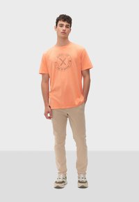 T-shirt arancione a maniche corte con un logo grafico rotondo al centro, abbinato a pantaloni beige e sneakers con dettagli verdi.