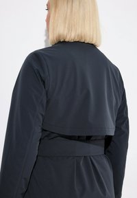 Giacca leggera nera con un design posteriore a strati, vita cintata e texture liscia, caratterizzata da una leggera lucentezza.