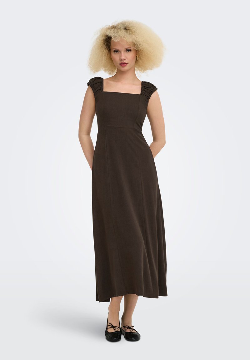 Femme aux cheveux blonds bouclés portant une robe midi marron foncé sans manches et des ballerines noires, debout sur un fond uni.