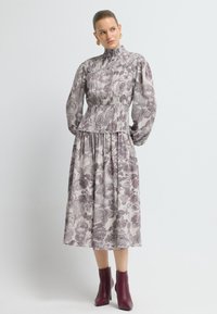 Largo vestido floral en tonos suaves de púrpura y crema, con un cuello alto, mangas abullonadas y una cintura ajustada, combinado con botas de tobillo burdeos.