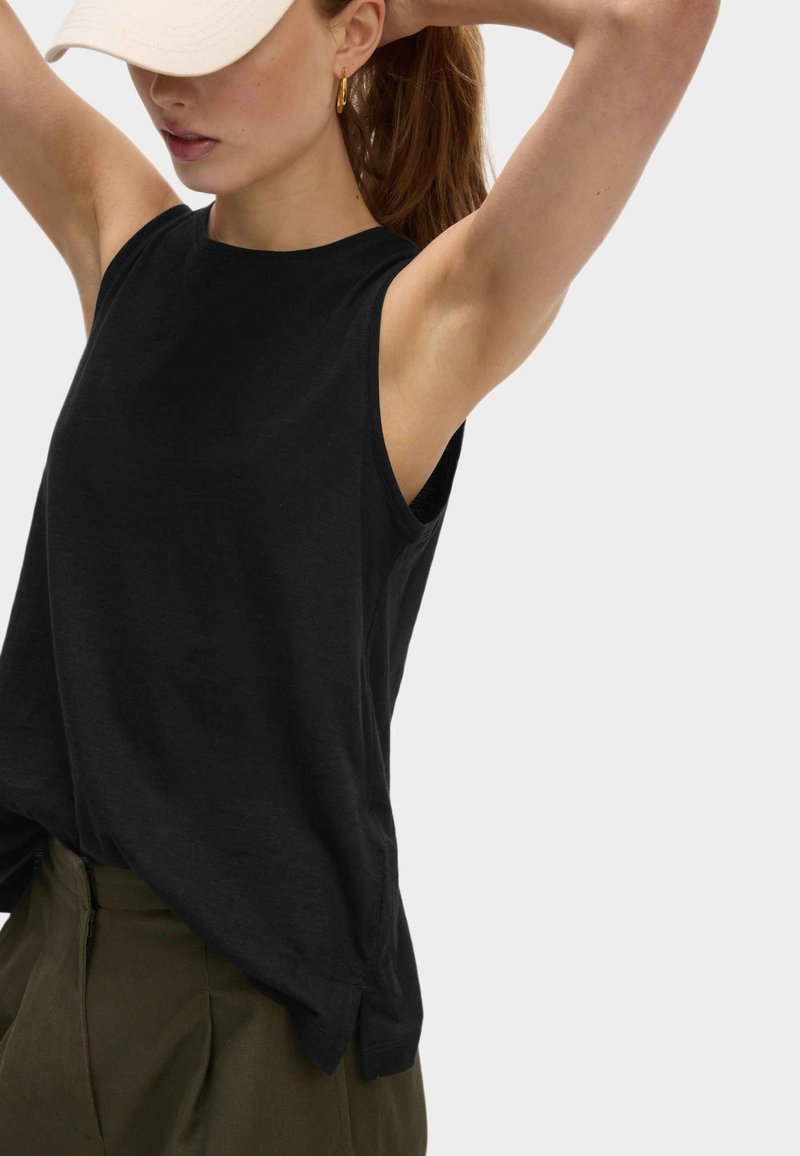 Mujer con una blusa negra sin mangas, pantalones verde oliva, una gorra beige y pendientes de aro dorados, ajustándose el cabello frente a un fondo liso.