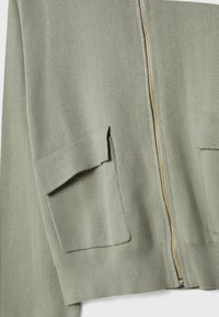 Gros plan d'un cardigan tricoté vert sauge avec fermeture éclair métallique et deux poches avant à rabat.