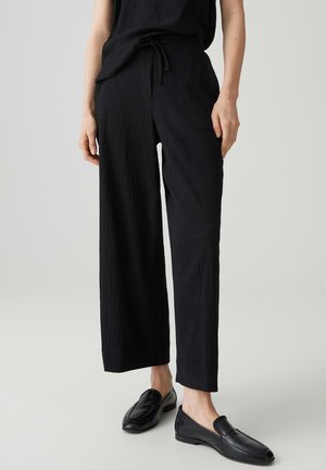 Person trägt schwarze, strukturierte, weit geschnittene Hose und schwarze Leder-Loafer, steht vor einem schlichten hellen Hintergrund.