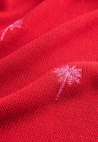 Červená pletená látka s malými svetloružovými vzormi palmových stromov, vtkanými do textúry, zobrazená v detailnom zábere.