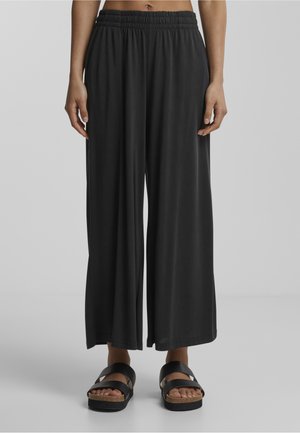 Femme portant un pantalon noir à jambes larges avec une taille élastique et des sandales noires, debout devant un fond gris clair uni.