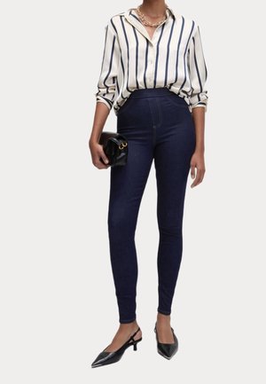 Witte en marineblauwe gestreepte blouse, high-waisted donkerblauwe jeans, zwarte handtas, zwarte puntige hakken en een gouden kettinghanger.