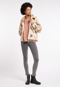 Beige Fleecejacke mit abstrakten Mustern, kombiniert mit einem rosa Hemd und grauen Jeans, ergänzt durch schwarze Ankle Boots.