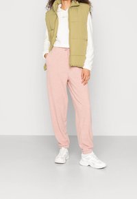 Licht olijfgroene gewatteerde vest over een witte, gestructureerde trui, gecombineerd met zachte roze joggers. Model draagt witte sneakers en staat op een neutrale achtergrond.