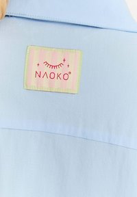 Svetlo modrá košeľa s béžovo-ružovým prúžkovaným záplatou s nápisom "NAAOKO" a červeným dizajnom rias pri golieri.