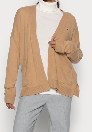 Personne portant un cardigan beige sur un col roulé blanc et un pantalon gris clair, tenant le cardigan ouvert de la main droite au niveau de la poitrine.