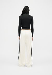 Γυναίκα που στέκεται με την πλάτη, φοράει μαύρο crop top με μακριά μανίκια και λευκά παντελόνια φαρδιά με μαύρες ρίγες στο πλάι.