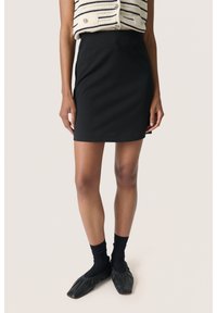 Zwarte rok met een aansluitende silhoutte, gemaakt van gladde stof. Gecombineerd met zwarte sokken en instap schoenen met geplooide randen.
