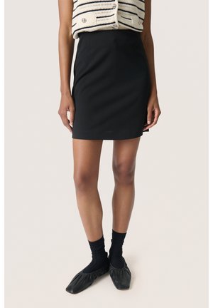 Jupe noire avec une silhouette ajustée, faite de tissu lisse. Associée à des chaussettes noires et des chaussures slip-on avec des bords froncés.