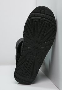 Semelle en caoutchouc noire avec un motif de bande de roulement texturé, présentant le logo UGG. La partie supérieure est douce et partiellement visible, montrant son matériau en laine.