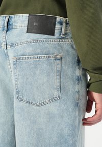 Lyseblå denimjeans med baklomme og svart merke, båret av en person i grønner langermet skjorte med hånden avslappet ved siden av.