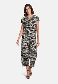 Zwart-witte jumpsuit met abstracte geel-groene vlekken, voorzien van een kraag, korte mouwen en wijde pijpen. Zwarte sandaaltjes met bandjes.