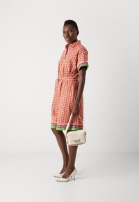 Kurzes Kleid mit kurzen Ärmeln in Orange mit Obstmuster, elastischem Bund, grünen Akzenten, kombiniert mit einer leichten Handtasche und beigen hohen Schuhen.