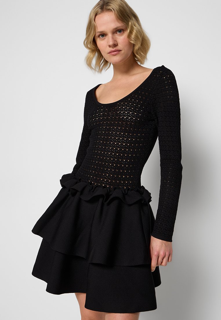 Schwarzes Kleid mit langen Ärmeln, das mit einem strukturierten Häkeloberteil und einem schichtweisen Rüschensockel versehen ist. Glattes Material und tailliertes Design.