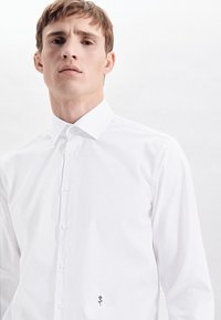 Witte button-up overhemd met een klassieke kraag, voorzien van een kleine donkere bloemborduring bij de zoom. Gladde katoen stof met een getailleerd ontwerp.