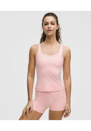 Kobieta stojąca, ubrana w jasnoróżowy dopasowany top na ramiączkach i pasujące szorty, z ręką opartą na udzie na jednolitym tle.