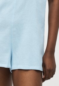 Κοντινό πλάνο ενός ατόμου που φοράει ανοιχτό μπλε υφασμάτινο σορτς, εμφανίζοντας τη λεπτομέρεια του υφάσματος και μέρος του δεξιού βραχίονα και χεριού.