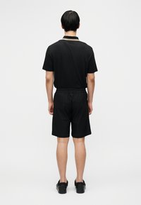 Μαύρο πόλο πουκάμισο με γιακά και χρυσές λεπτομέρειες, συνδυασμένο με μαύρο σορτς από δίχτυ. Το σύνολο ολοκληρώνεται με μαύρα υποδήματα.