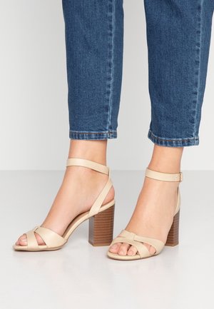 Beige blokhakken sandalen met gekruiste voorriemen en enkelbandje. Gelaagde bruine hak voegt visuele interesse toe. Draagt met cropped denim.