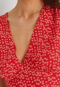 Abito floreale rosso con scollo a V profondo, dettagli arricciati e piccole fiori bianchi stampati su un tessuto morbido.