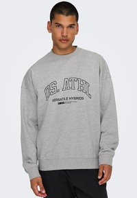 Gray sweatshirt with a crew neck, oversized fit, and black lettering "OS. ATHL VERSATILE HYBRIDS" printed across the chest. Cotton blend material.