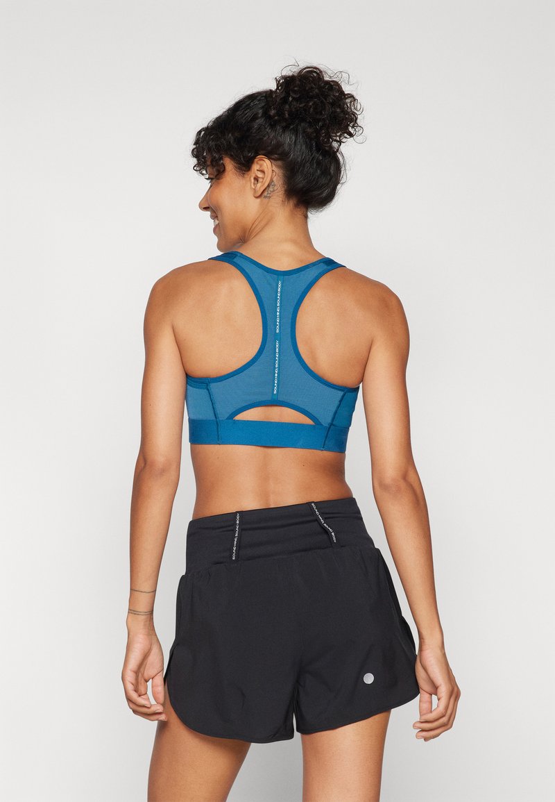 Sostén deportivo azul con detalle recortado y tirantes ajustables, combinado con pantalones cortos deportivos negros. Tejido suave, diseño de espalda de nadador, acentos minimalistas.