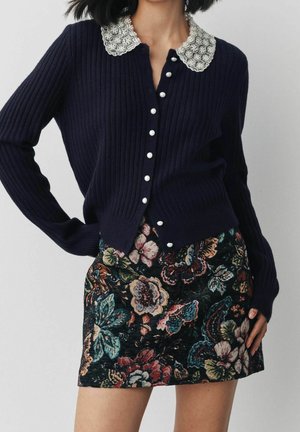 Femme portant un cardigan côtelé bleu marine avec des boutons en perles et un col brodé de perles, associé à une jupe colorée à motif floral.