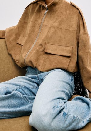 Persoon draagt een losse bruine suède jas met rits en grote zakken, gecombineerd met lichtblauwe wijde jeans, zittend op een bruine bank.