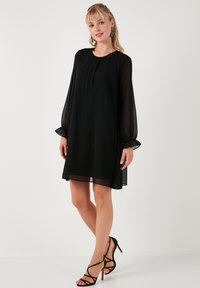 Black shift dress with long sheer sleeves and a round neckline. Soft fabric, pleats at the front, and above-the-knee length.