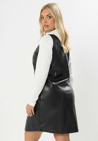 Abito in ecopelle nera con design senza maniche, caratterizzato da una scollatura a V e una texture liscia, sovrapposto a una maglietta bianca a maniche lunghe.