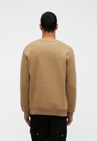 Férfi rövid fekete hajjal, háttal állva, bézs pulóverben és fekete munkásnadrágban egy sima fehér háttér előtt.