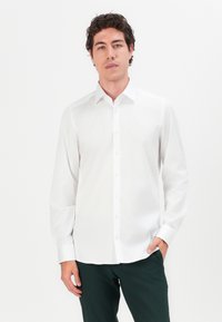 Weißes Langarmhemd mit Button-Down-Kragen, Vorderleiste und Ein-Knopf-Manschetten, kombiniert mit dunkelgrüner Hose.