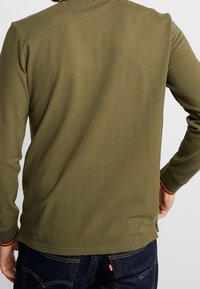 Oliwkowa koszula z długim rękawem, wykonana z bawełny, z okrągłym dekoltem i kontrastowymi pomarańczowymi detalami na kołnierzyku i mankietach.