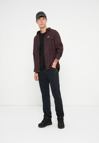 Camicia di flanella a scacchi rossi e neri sopra una t-shirt nera, abbinata a jeans scuri e sneakers nere. Il modello è in piedi con una mano in tasca.