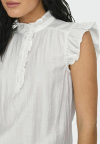 Blusa sin mangas blanca con cuello y aberturas de los brazos con volantes, que presenta una tela texturizada con sutiles líneas verticales. Se aprecia un cierre de botón.